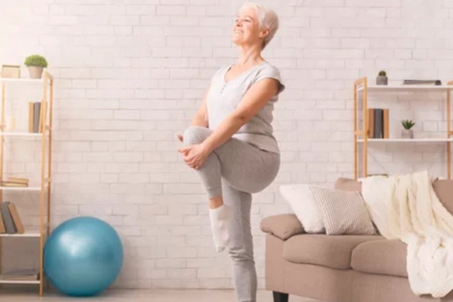 An older adult practising a standing balance exercise with light support from a chair.
